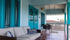 A covered porch with a wicker sofa, teal shutters, and a view of the ocean.