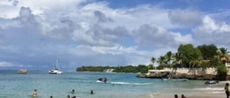 A beach with clear water, people swimming and sunbathing, boats, and a distant island with palm trees.