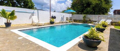 Une piscine rectangulaire aux bords blancs, entourée de chaises longues blanches et de plantes en pot.