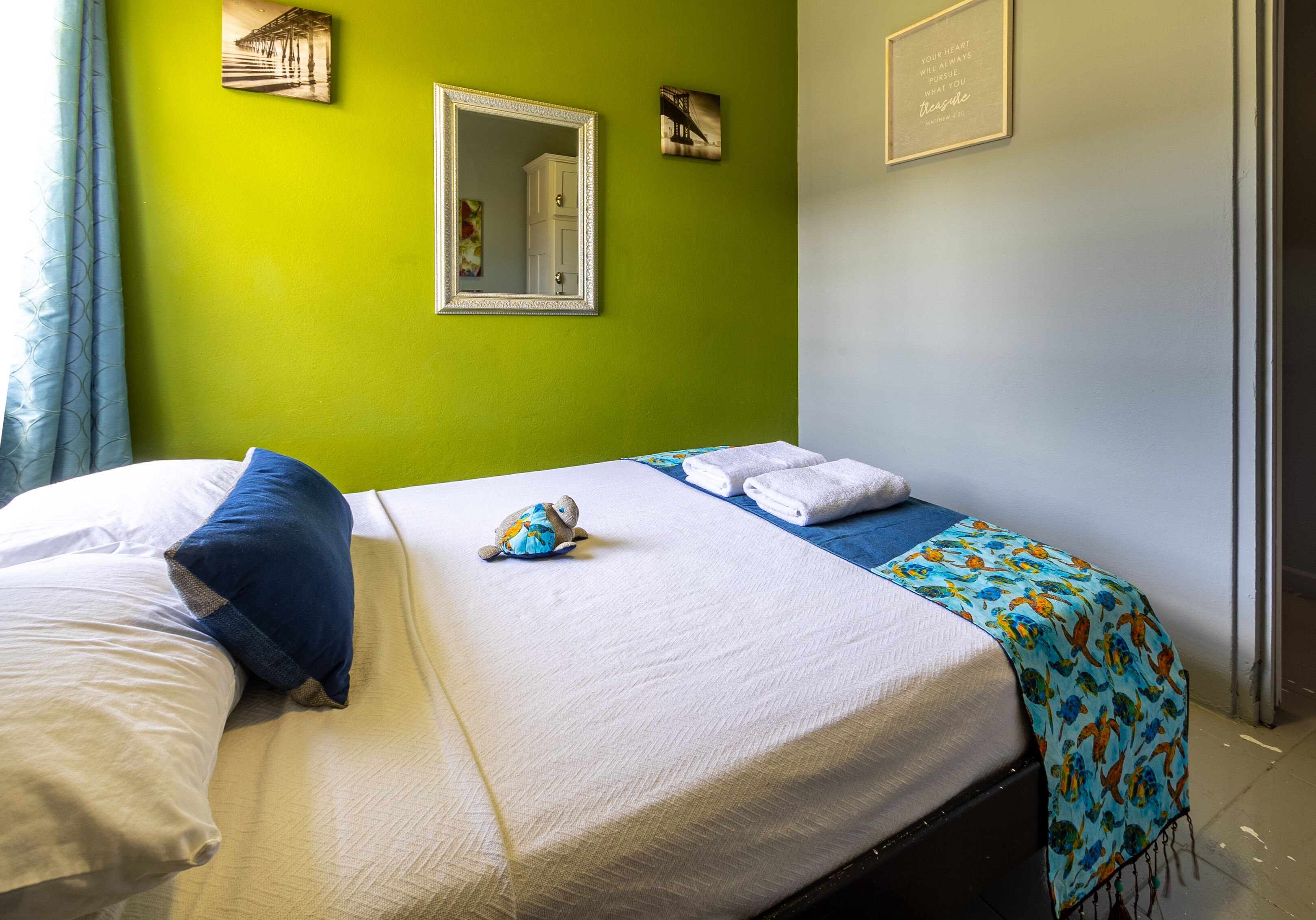 A neatly made bed with white linens and a blue pillow, a blue blanket with a pattern, and a small turtle plush toy on the bed.