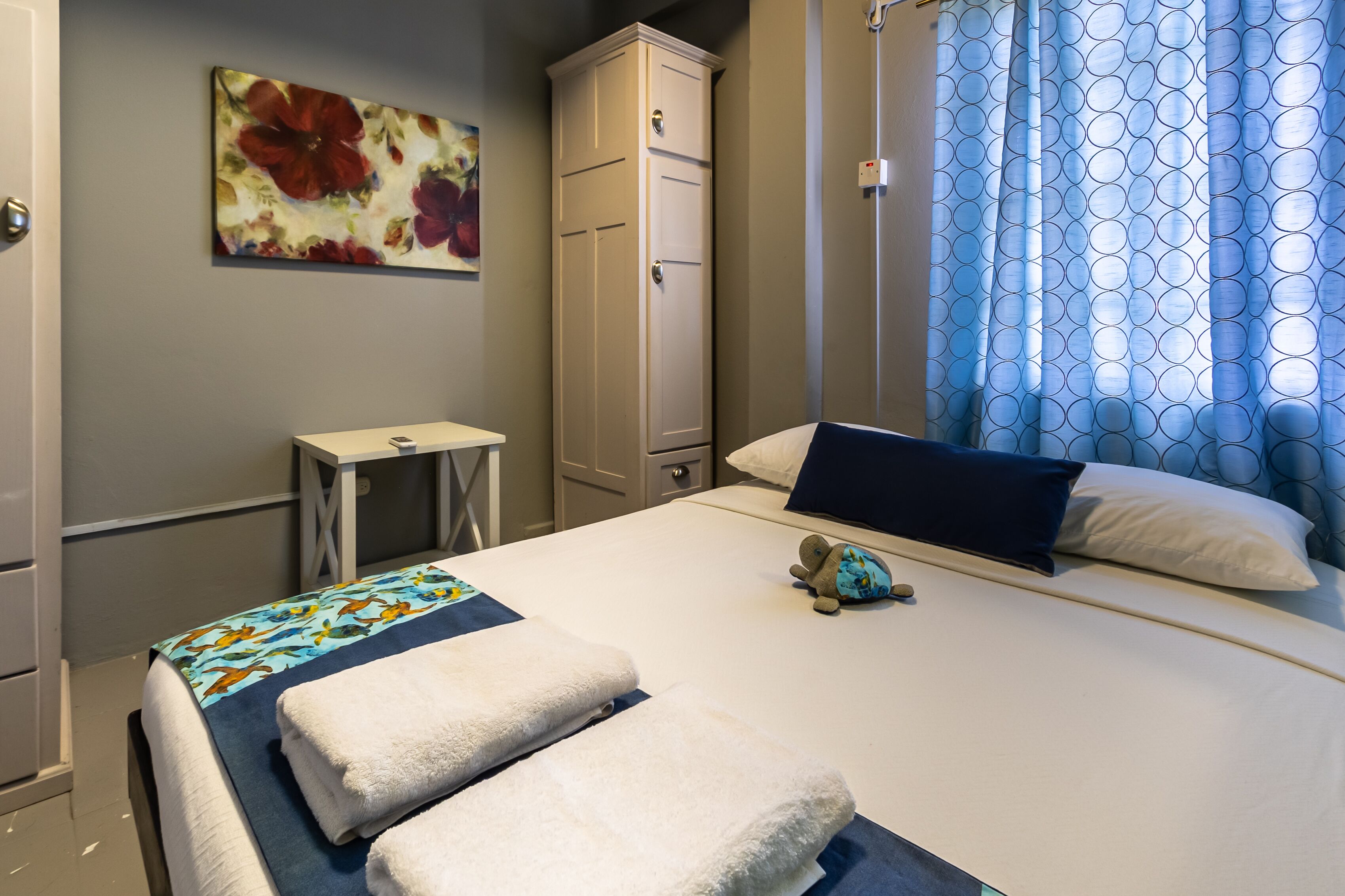 A hotel room with a bed, a small table, a wardrobe, a painting, and a window with patterned curtains.