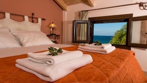 A bedroom with a bed, a window with a view of the sea, and a nightstand with a lamp. - Argo Maisonettes & Suites (Lefkada)