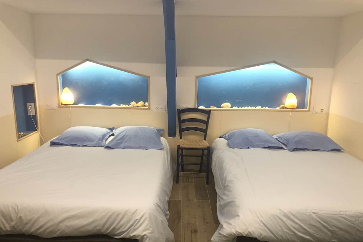 Ein Zimmer mit zwei Betten, jeweils mit Kopfteil und Fenster. Eine Sitzgelegenheit steht zwischen den Betten, und ein kleiner Tisch mit einer Lampe ist ebenfalls vorhanden.