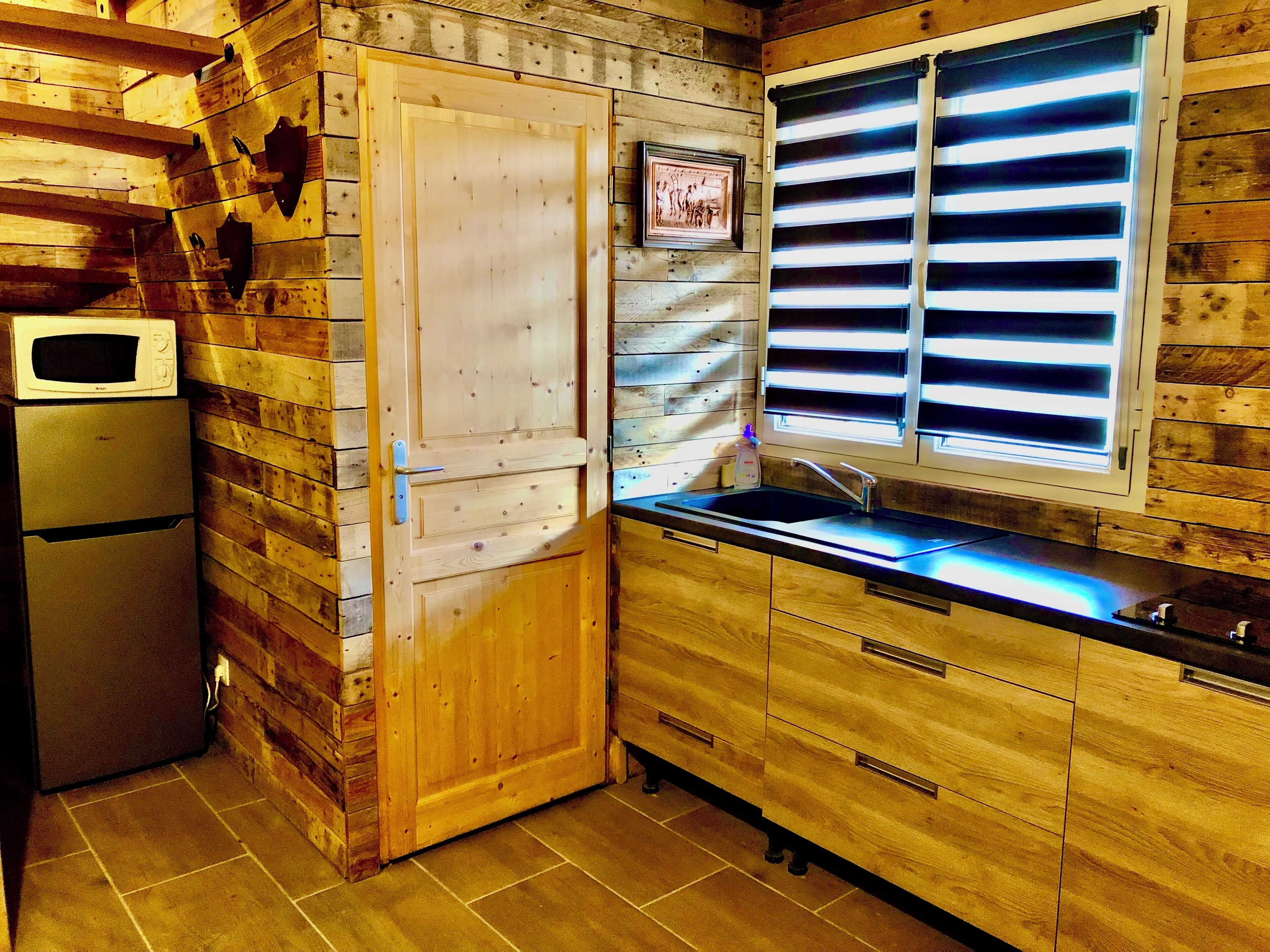 Una cocina con paredes de madera, un refrigerador, un microondas, un lavamanos y una ventana con persianas.