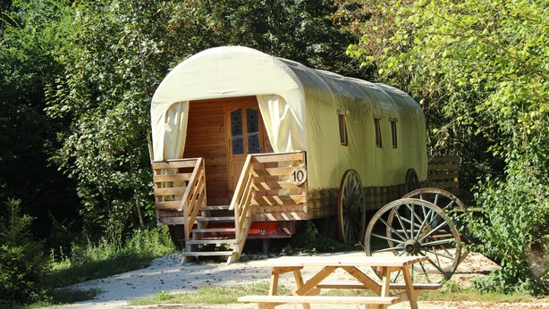 Ein Wohnmobil mit Veranda, einer Holztür und Stufen, ein Picknicktisch davor und ein großes Rad im Blickwinkel.