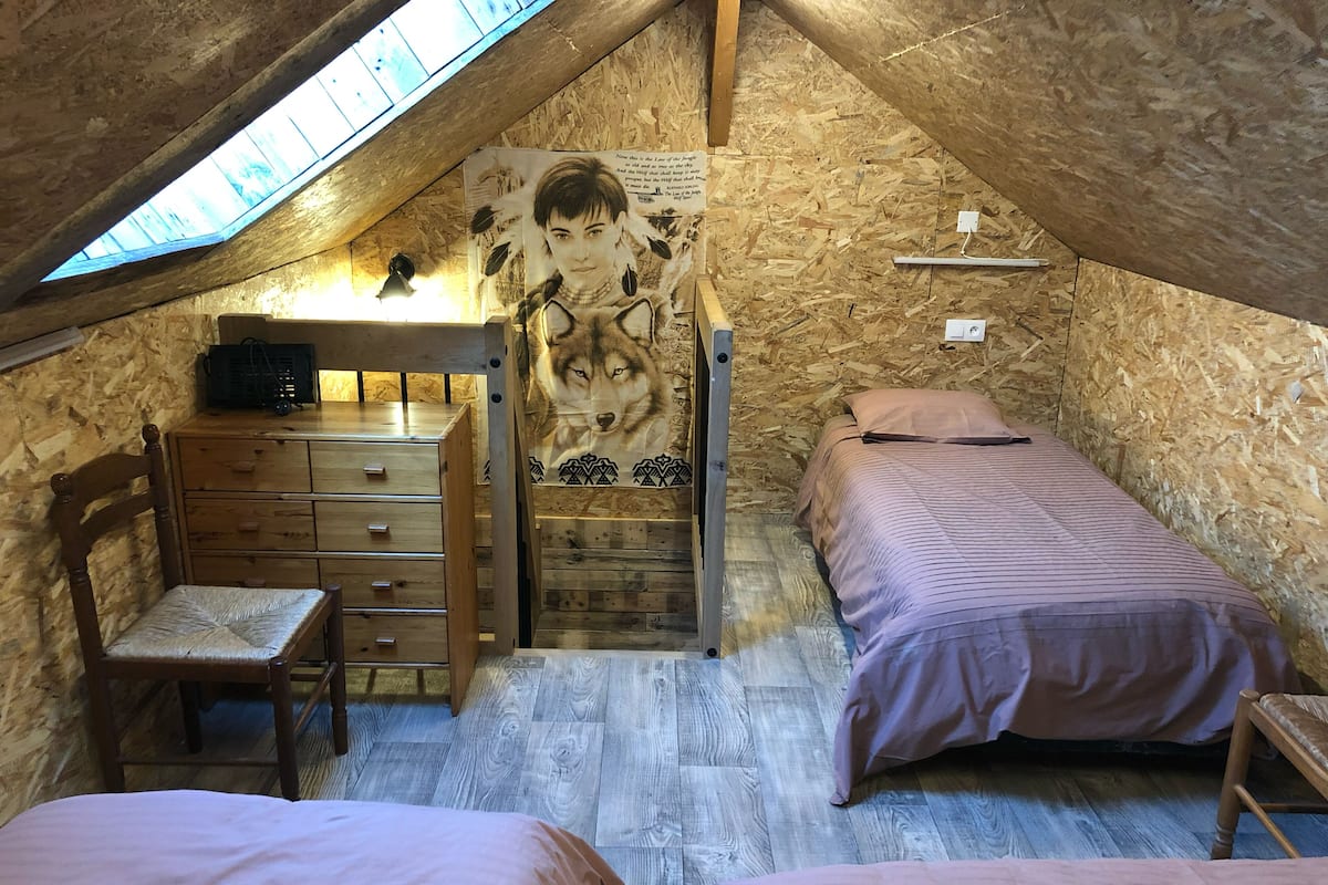 Ein gemütliches Schlafzimmer im Dachgeschoss mit schrägen Wänden, Holzwänden, einem Schreibtisch mit Schubladen, einem Stuhl, zwei Betten mit rosa Bettwäsche und einem Wolfsposter.