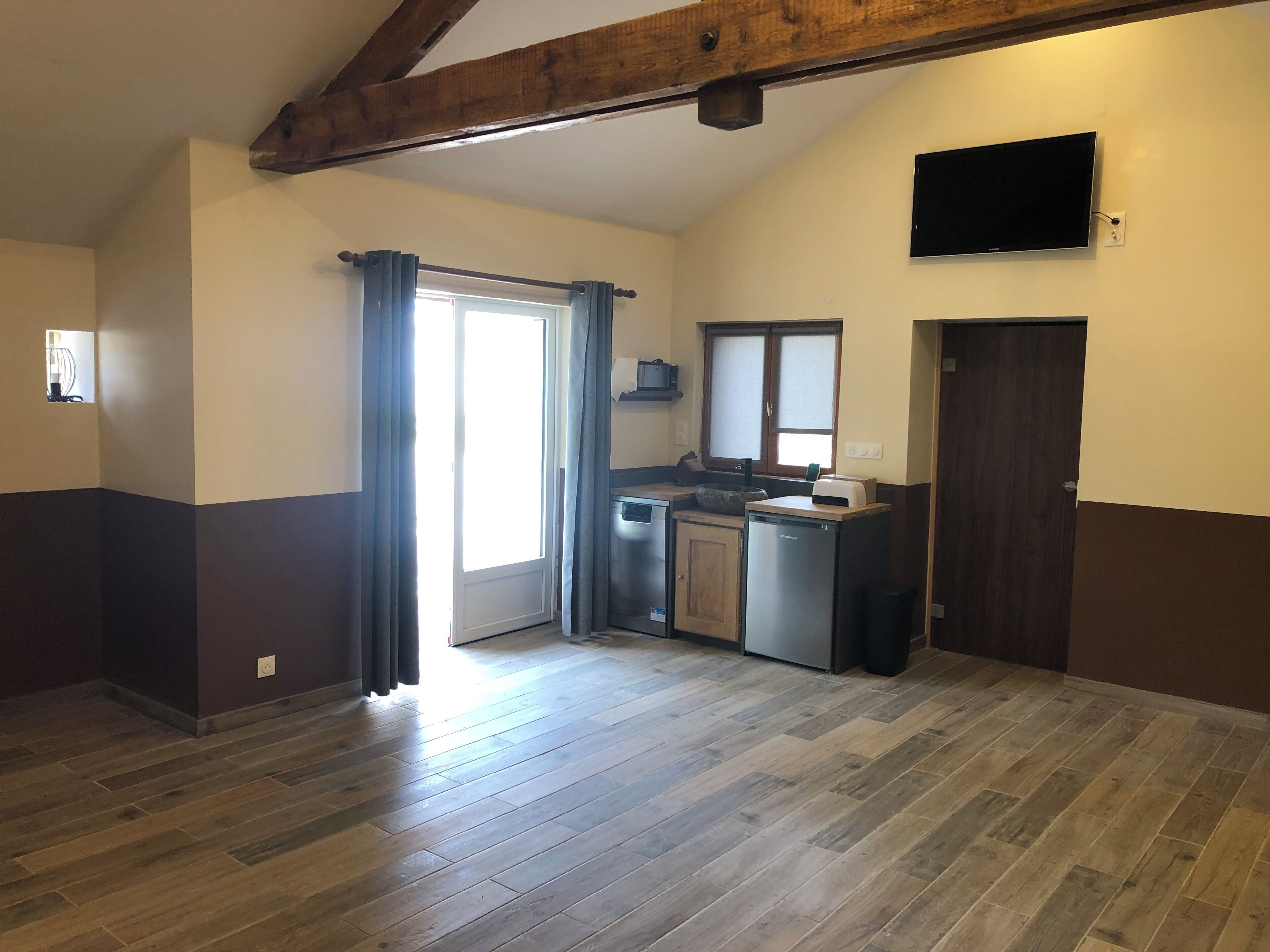 A kitchen with a built-in dishwasher, a microwave, and a sink. There is a window with curtains, a door, and a television mounted on the wall.