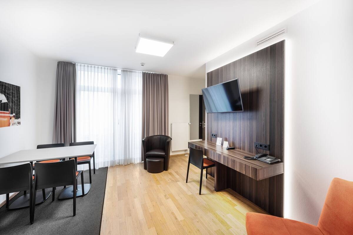 A modern hotel room with a wooden desk, a flat-screen TV mounted on the wall, a dining area with a table and chairs, and a large window with curtains.