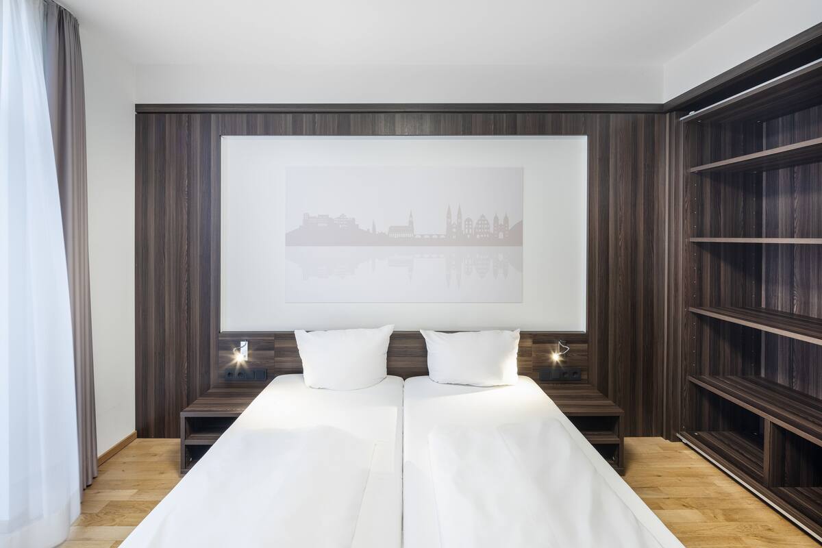 A modern hotel room with a large bed, a cityscape artwork on the wall, and a built-in bookshelf.