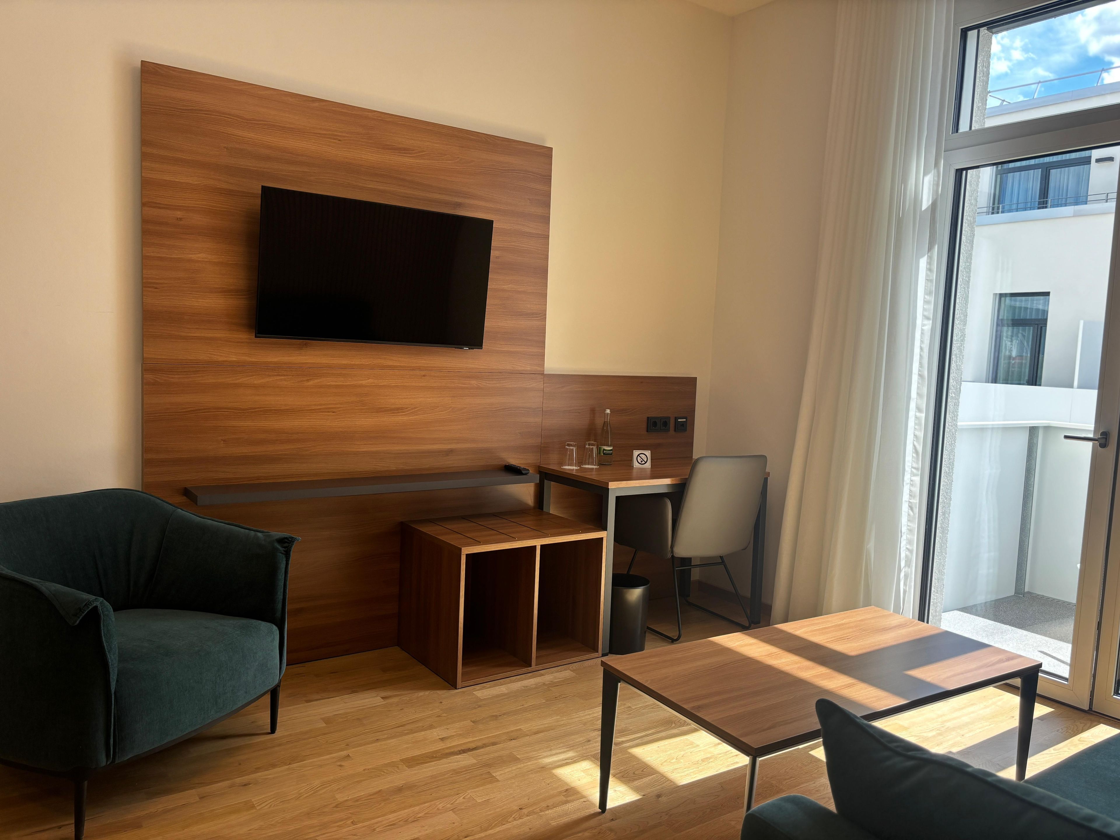 Una habitación de hotel moderna con un televisor de pantalla plana montado sobre un panel de madera, un escritorio de madera pequeño con una silla, un sofá y una puerta de cristal que da a un balcón.
