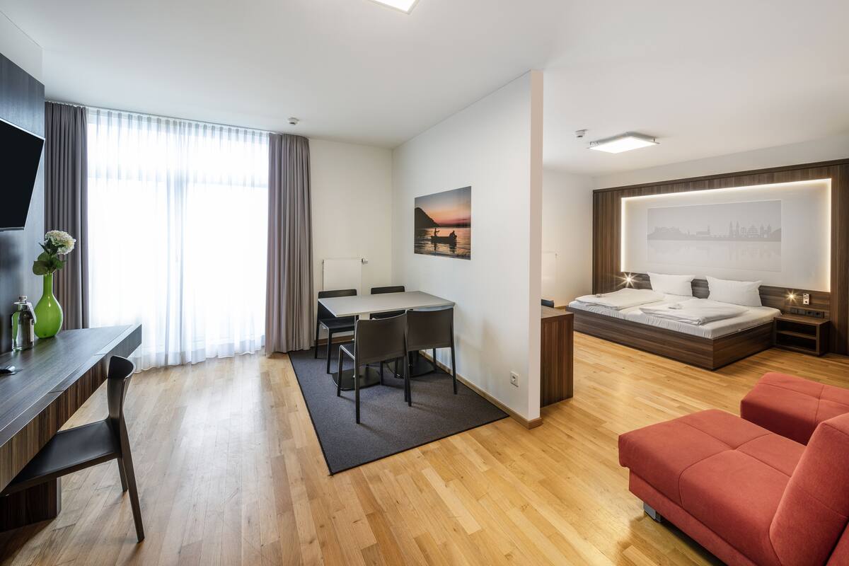 A modern hotel room with a wooden floor, a dining area with a table and chairs, a bed with a cityscape mural, and a red sofa.