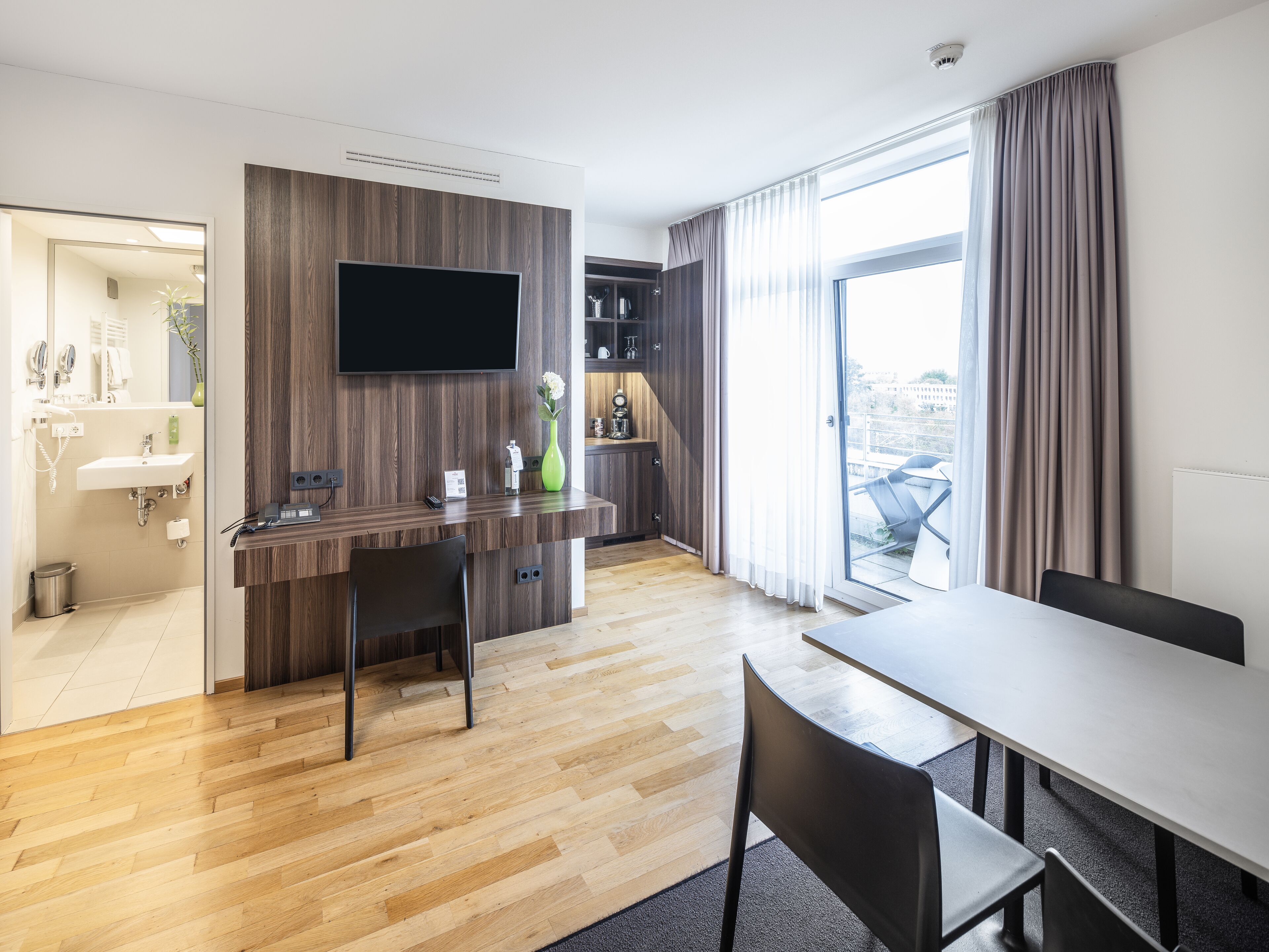 Une chambre d’hôtel moderne dotée d’un bureau en bois, d’un téléviseur à écran plat, d’un coin repas et d’une grande fenêtre avec des rideaux.
