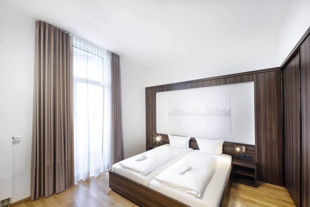 A modern hotel room with a large bed, wooden headboard, and a cityscape artwork on the wall.