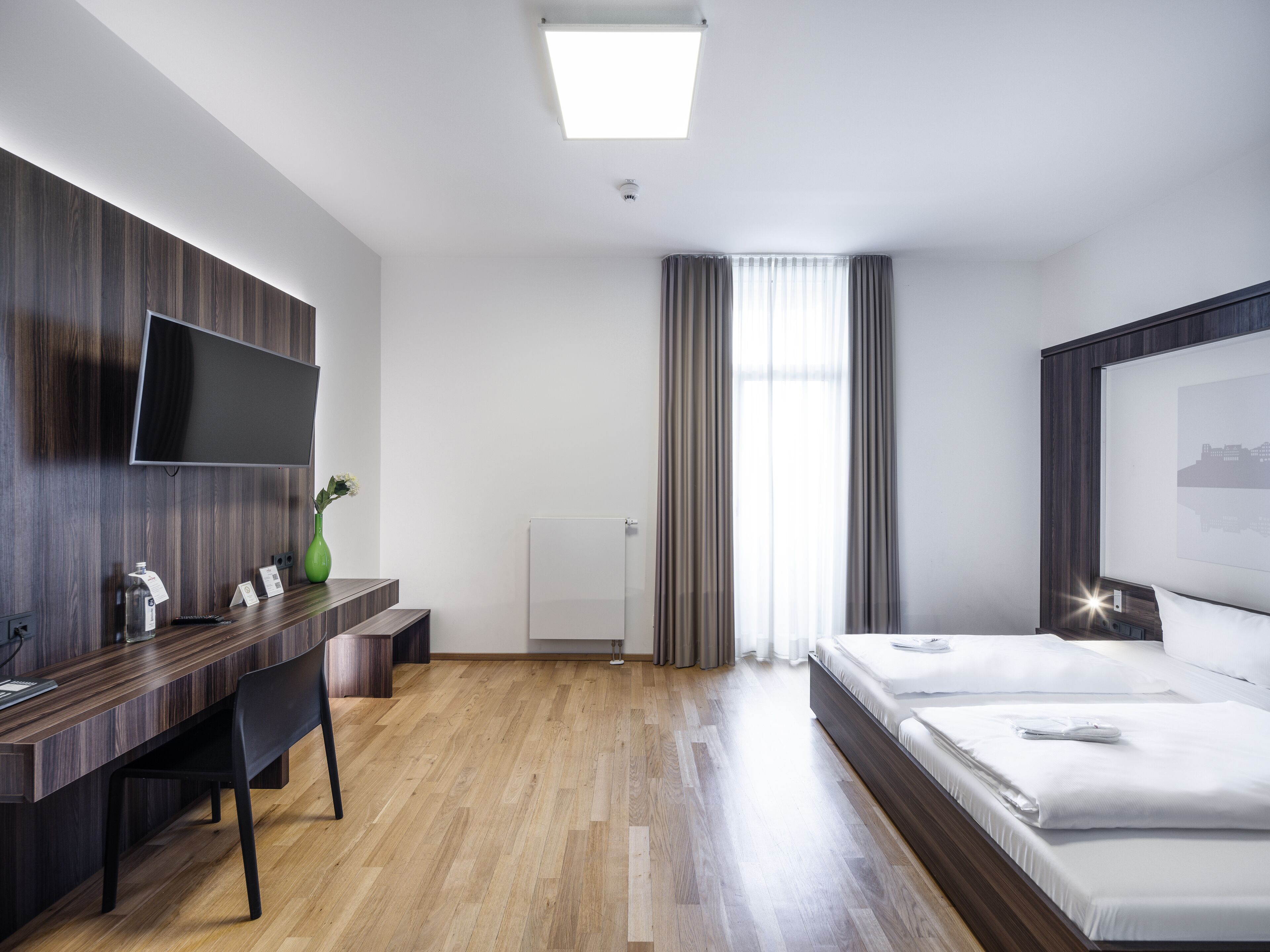A modern hotel room with a wooden desk, a flat-screen TV, a bed, and a large window with curtains.