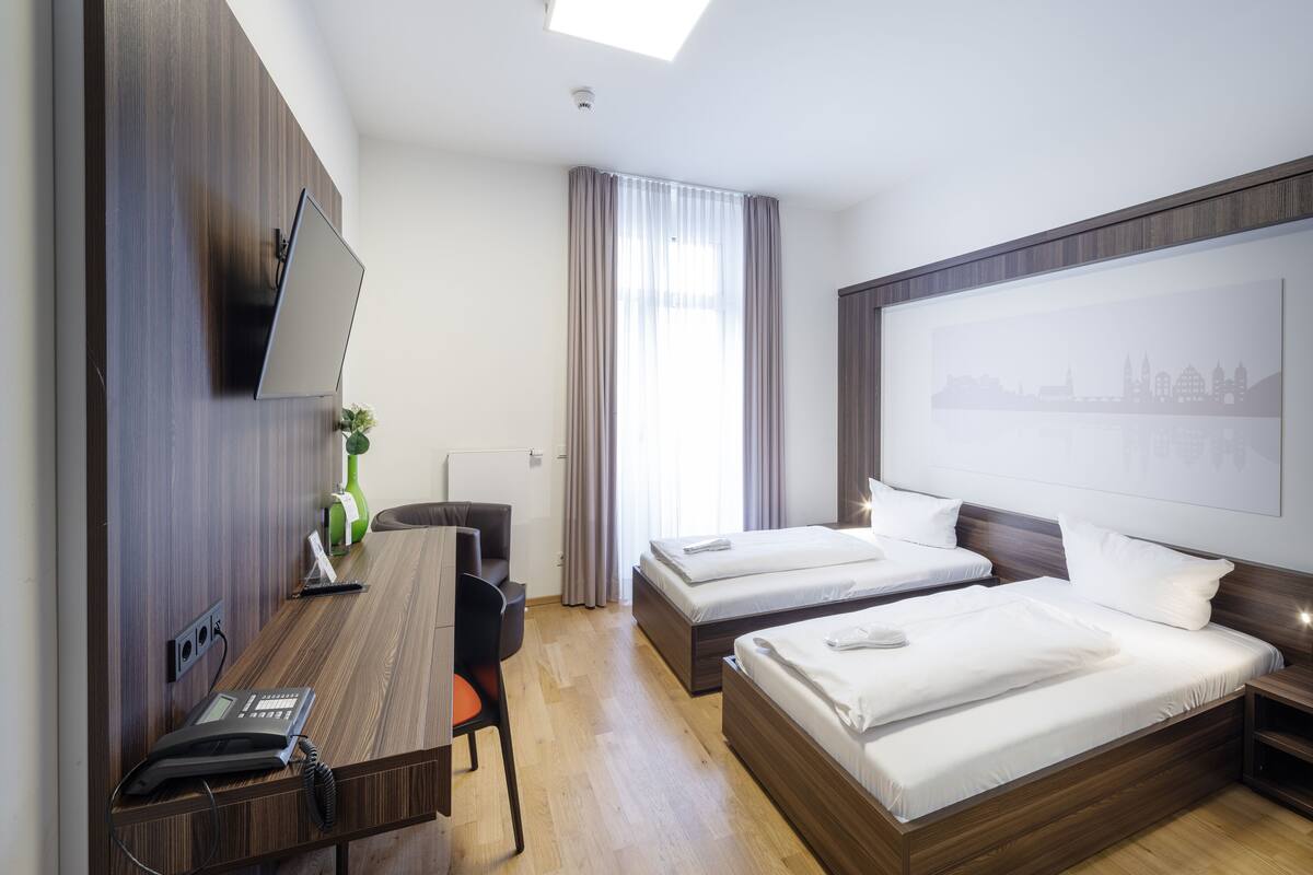 A hotel room with two beds, a desk with a telephone, a flat-screen TV mounted on the wall, and a cityscape artwork above the beds.