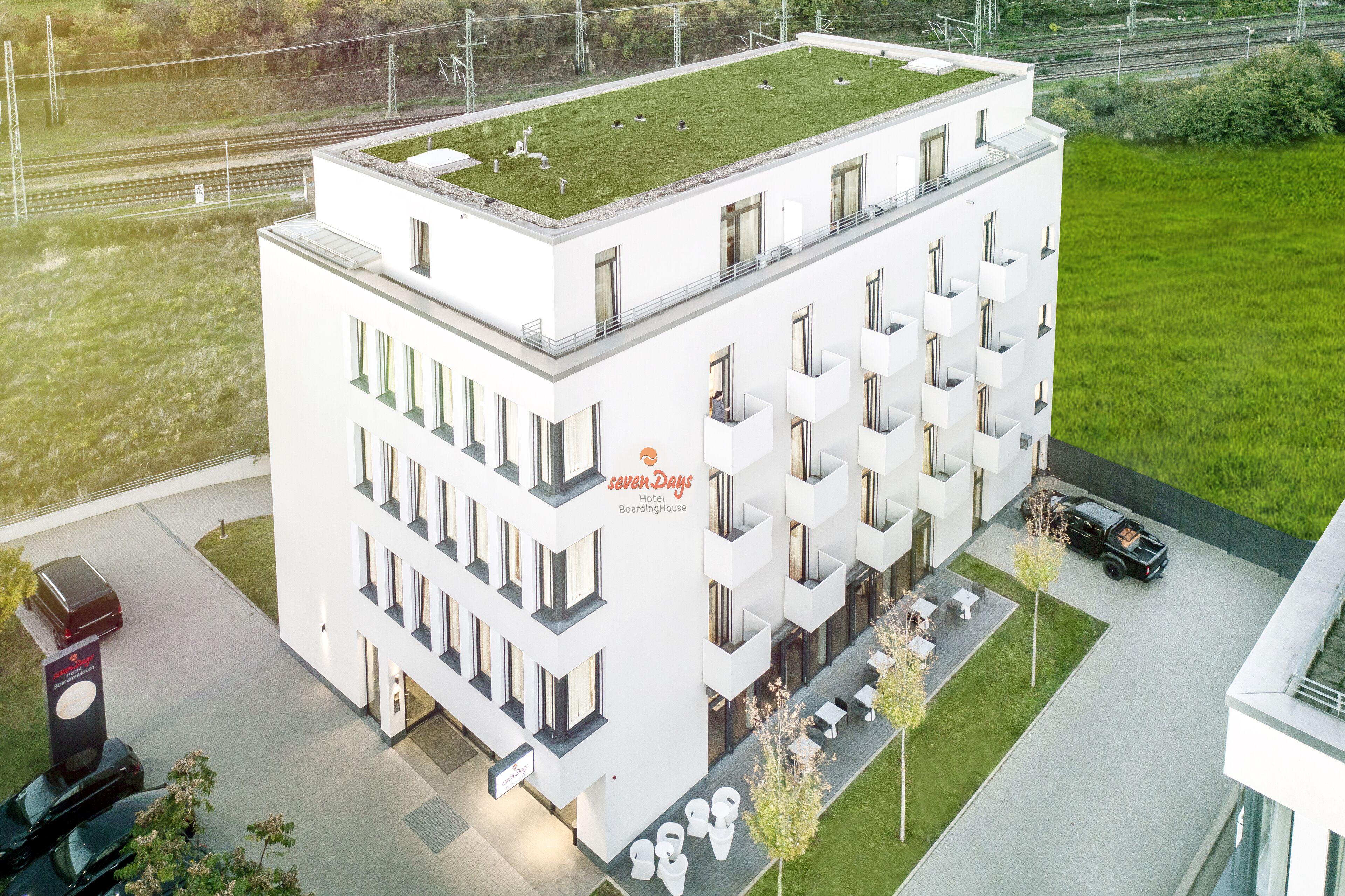 A modern hotel with a flat roof, multiple balconies, and a parking area.