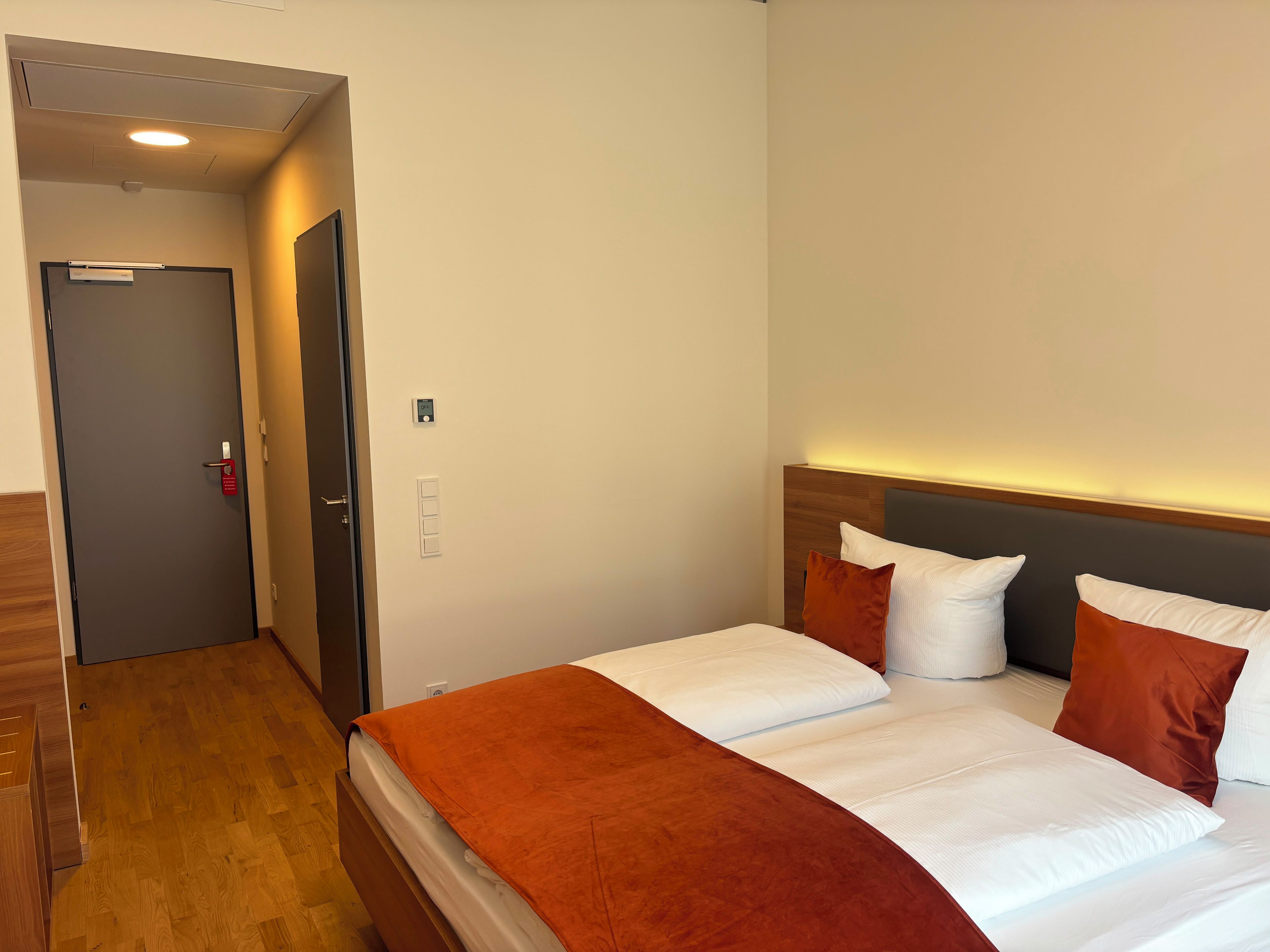 Hotellihuone, jossa on sänky, ovi ja seinässä oleva valokytkin.