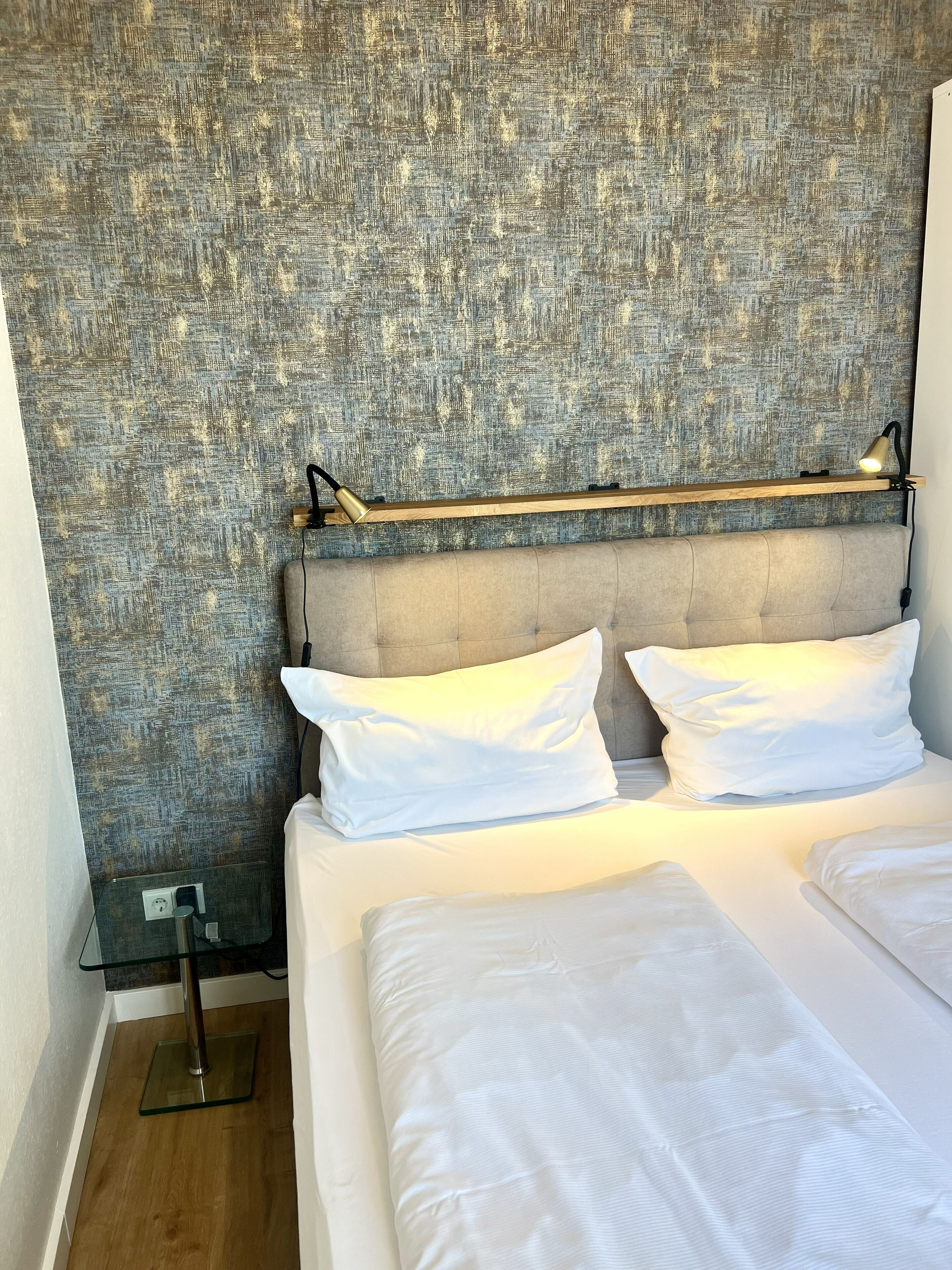 A hotel room with a textured wall, a grey upholstered headboard, two white pillows, a bedside table with a lamp, and a wooden floor.