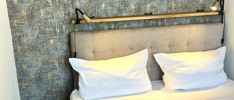 A hotel room with a textured wall, a grey upholstered headboard, two white pillows, a bedside table with a lamp, and a wooden floor.