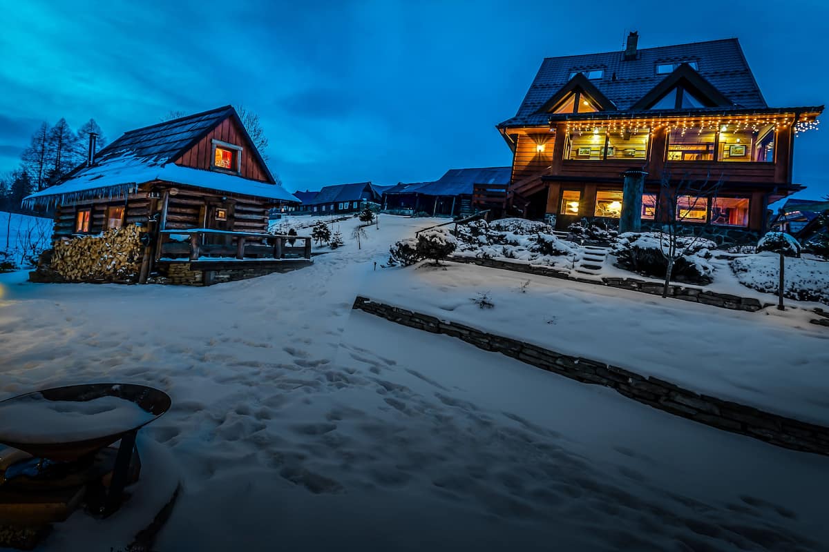 Una cabaña de troncos con un camino de piedra que lleva a la entrada, rodeada de terreno cubierto de nieve.