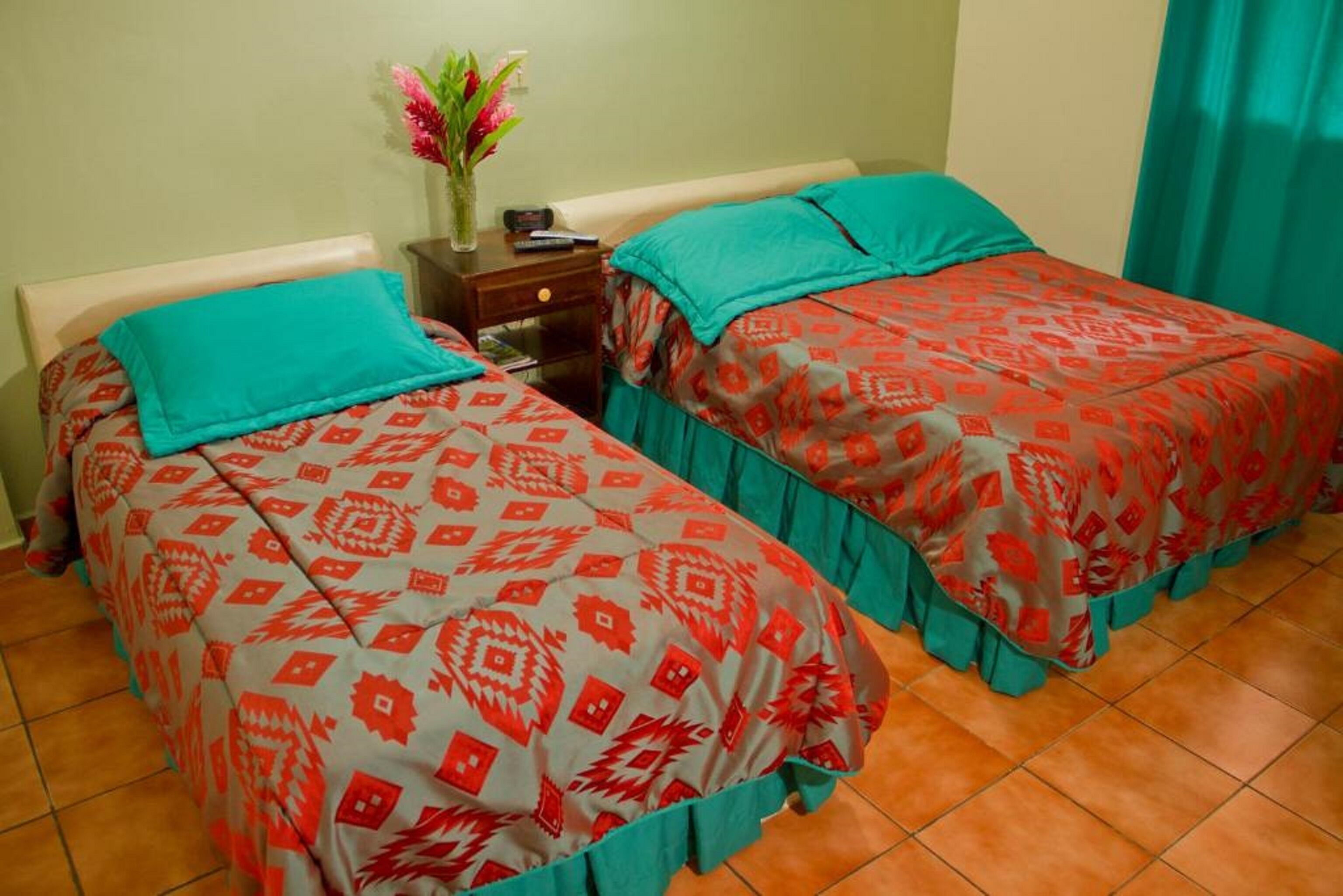 Habitación de hotel con dos camas, cada una con colcha estampada y almohadas color turquesa. Se observa un mesita de noche con un florero y un control remoto.