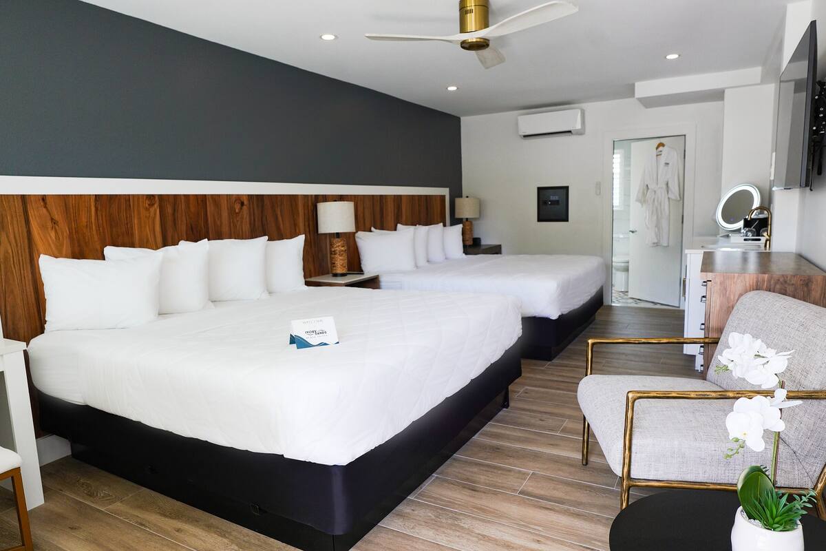 Quarto de hotel com duas camas, uma escrivaninha, uma cadeira, uma pequena mesa com uma planta e um banheiro visível por uma porta aberta.