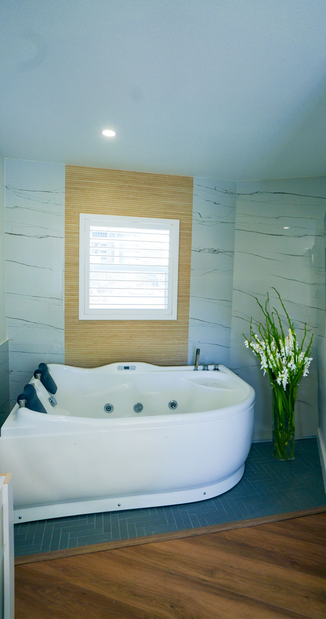 A modern bathroom with a freestanding bathtub, a window with blinds, and a vase with white flowers.