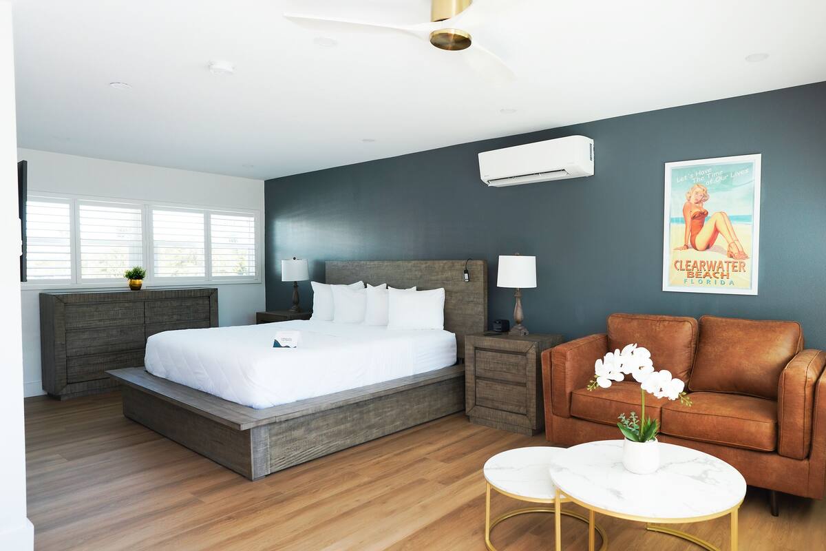 Quarto moderno com uma cama grande, mesinhas de cabeceira, um criado-mudo, um sofá de couro marrom, uma pequena mesa branca e um pôster na parede.