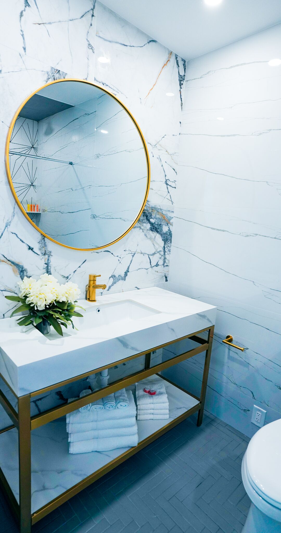 A bathroom with a marble wall, a round mirror with a gold frame, a white sink, and a gold vanity with towels.