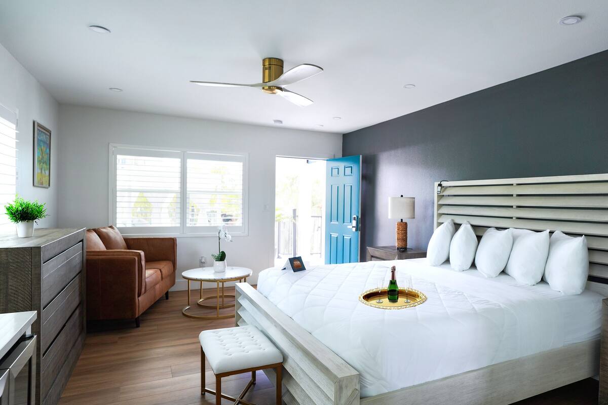 Um quarto moderno com uma cama grande, um ventilador de teto, uma mesa de centro, um sofá e uma mesinha de cabeceira com abajur.