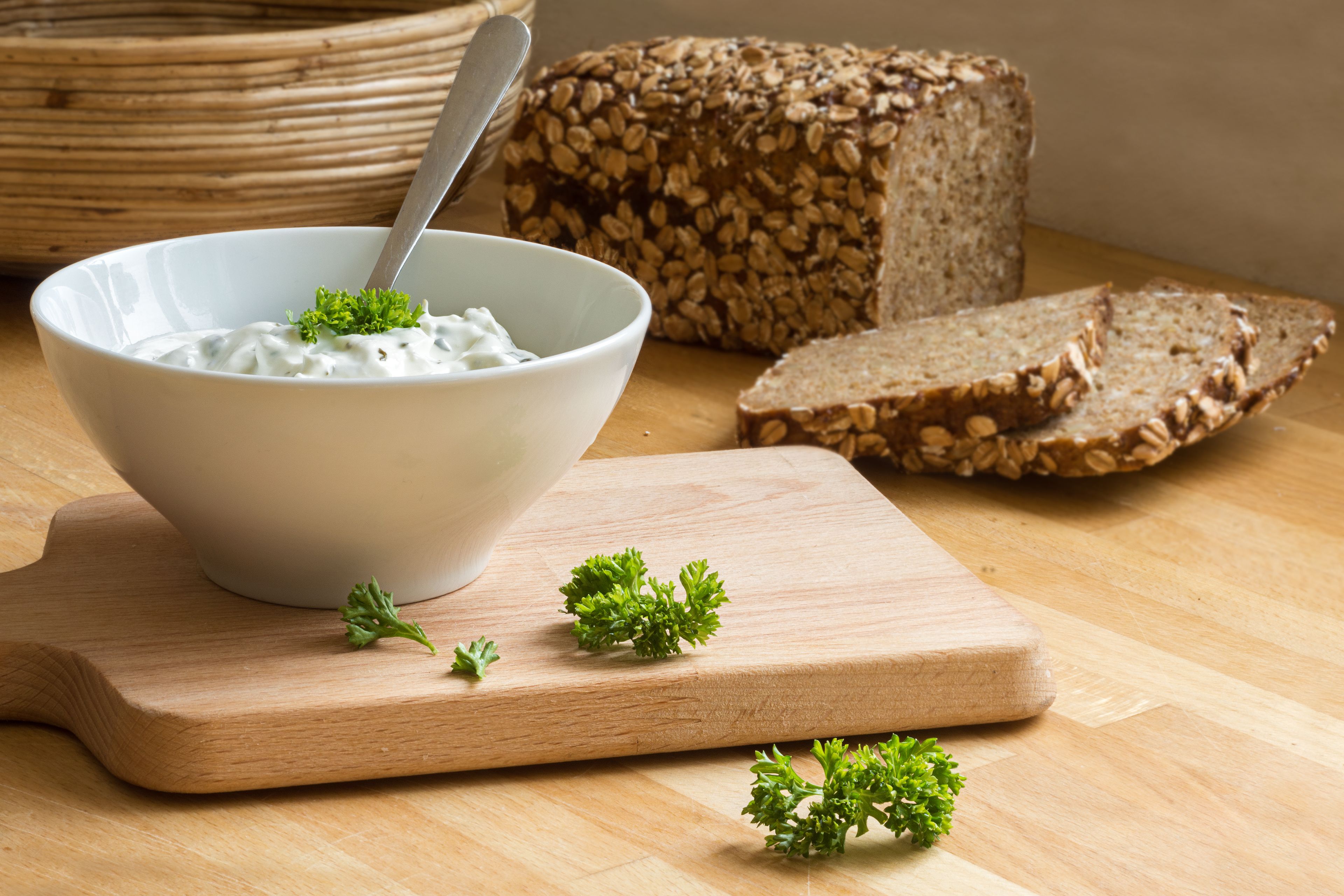 A bowl of yogurt with parsley on a wooden cutting board, slices of whole grain bread, and a loaf of bread in the background.