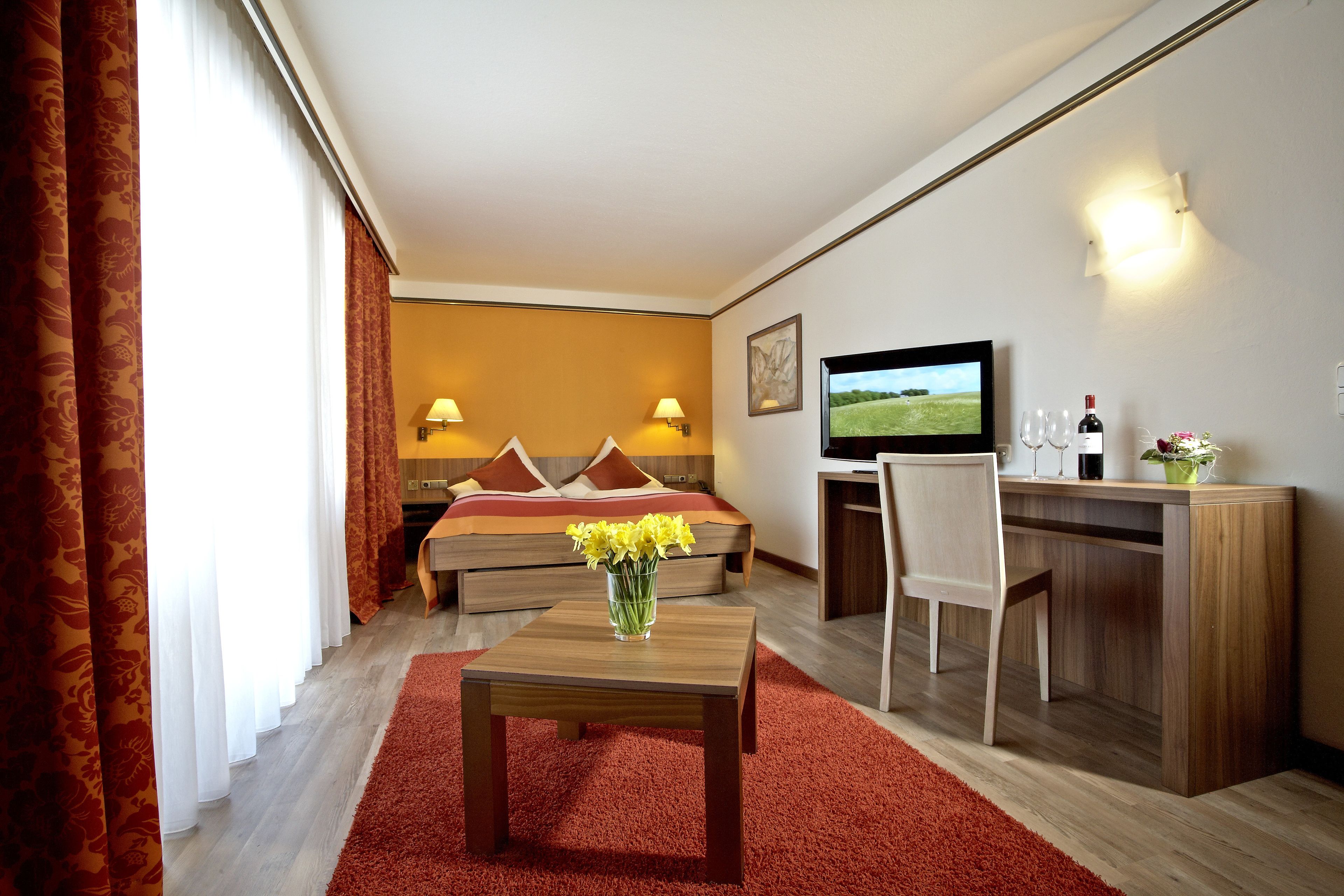 Une chambre d’hôtel avec un lit, un bureau et une chaise, une télévision et un vase avec des fleurs.