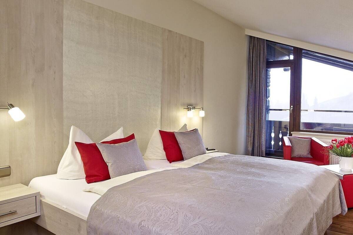 Une chambre d’hôtel avec un grand lit, deux tables de chevet, une petite table et une fenêtre avec des rideaux.