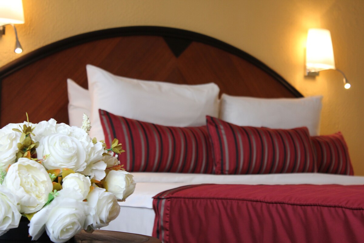 A hotel room with a wooden headboard, white bedding, red and striped pillows, and a vase of white flowers on the nightstand.