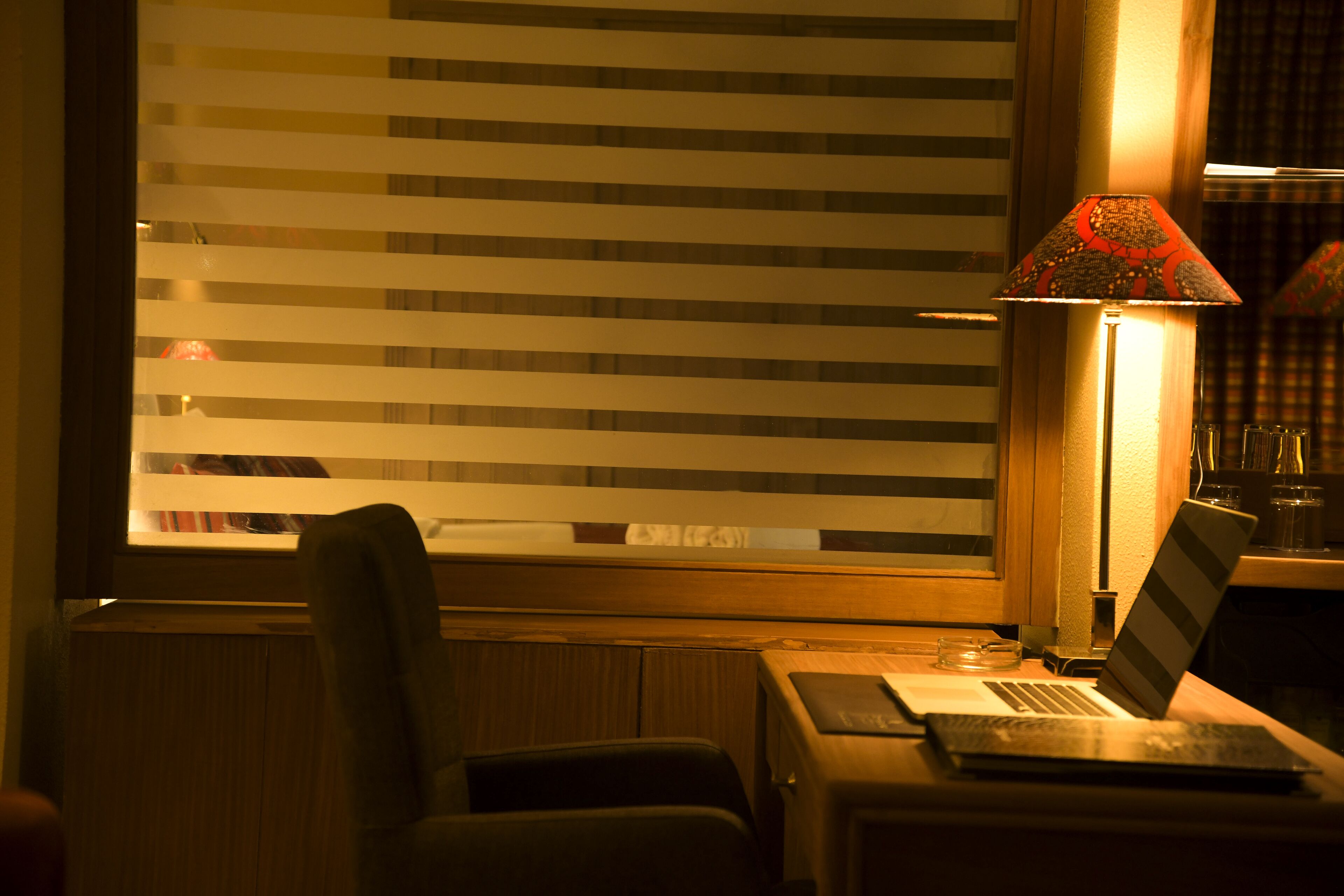 A room with a window with horizontal blinds, a desk with a laptop, a lamp, and a chair.