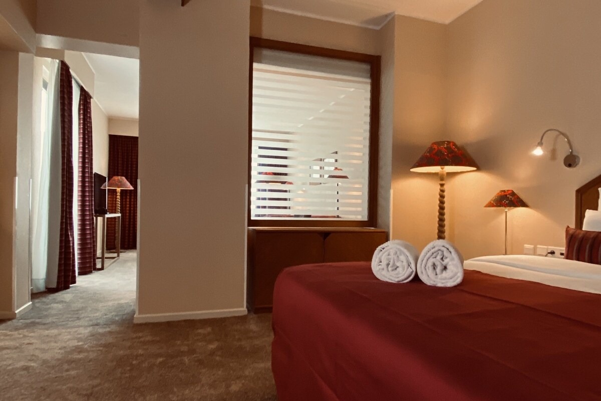 A hotel room with a bed, two lamps, a window with blinds, a carpeted floor, and a hallway with curtains.