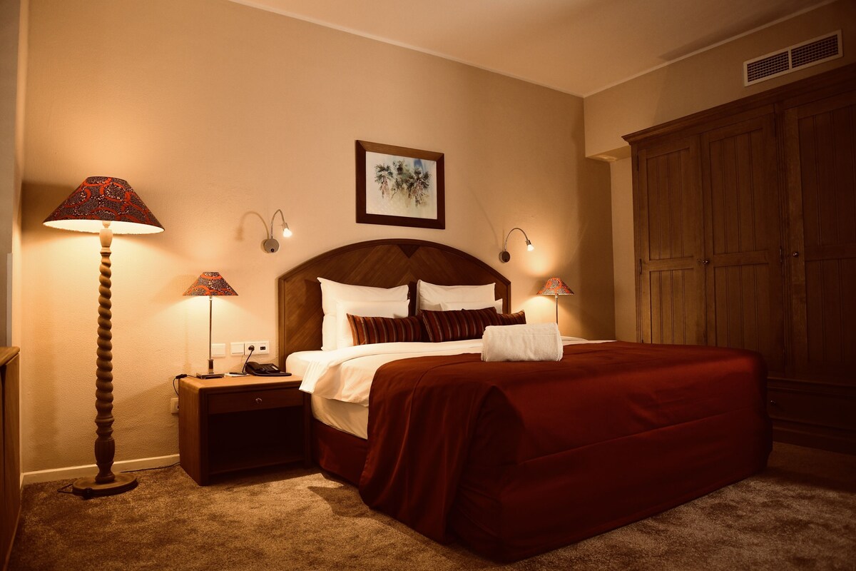 A neatly made bed with a red bedspread, two bedside lamps, a nightstand with a phone, and a wooden wardrobe.