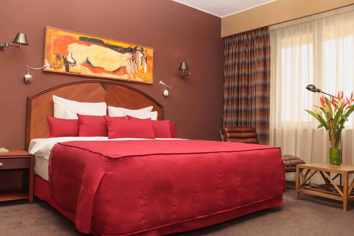 A hotel room with a large bed, a wooden headboard, red bedding, a painting above the bed, two wall-mounted lamps, a chair, a small table, and a window with curtains.