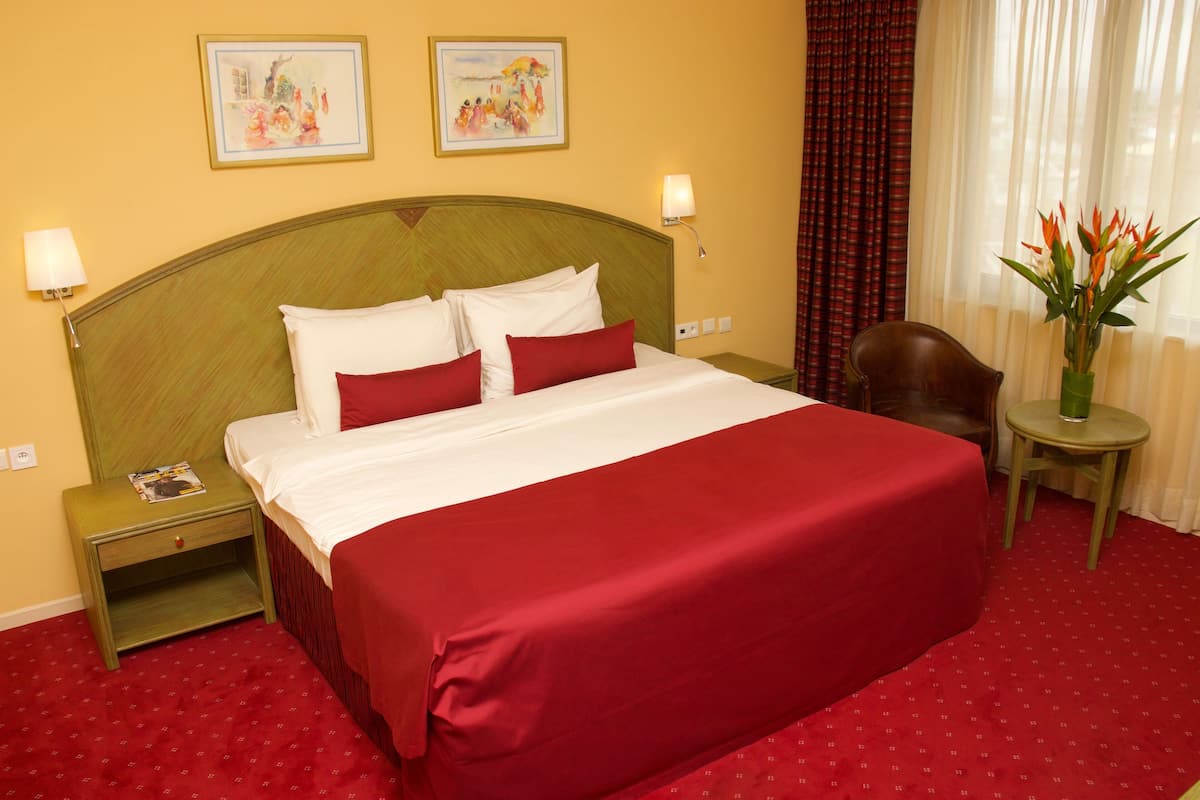 A hotel room with a large bed, red bedding, two wall-mounted lamps, a nightstand with a magazine, a chair, a small table with a vase, and two paintings on the wall.
