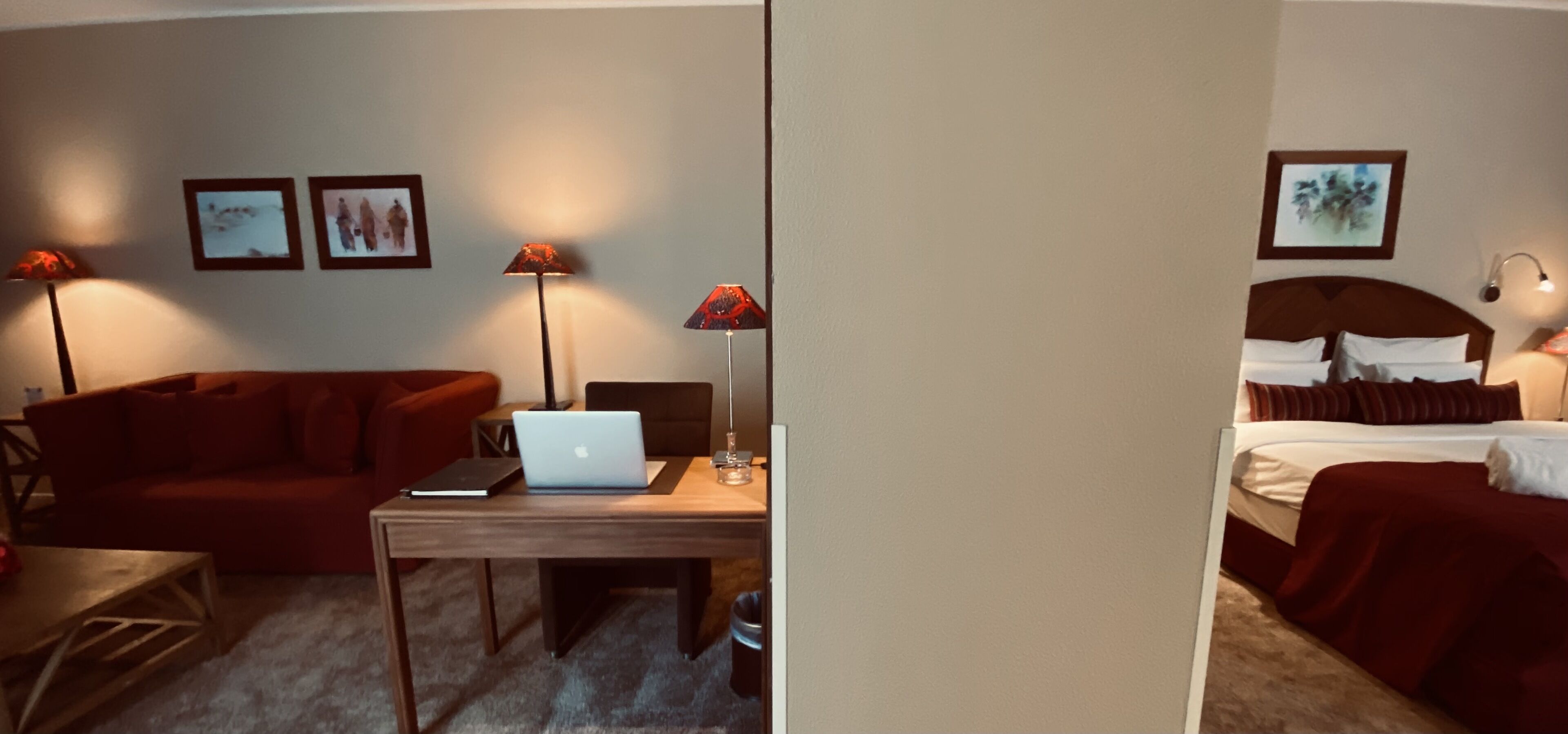A hotel room with a red sofa, a wooden desk with a laptop, two floor lamps, and a bed with a red blanket.