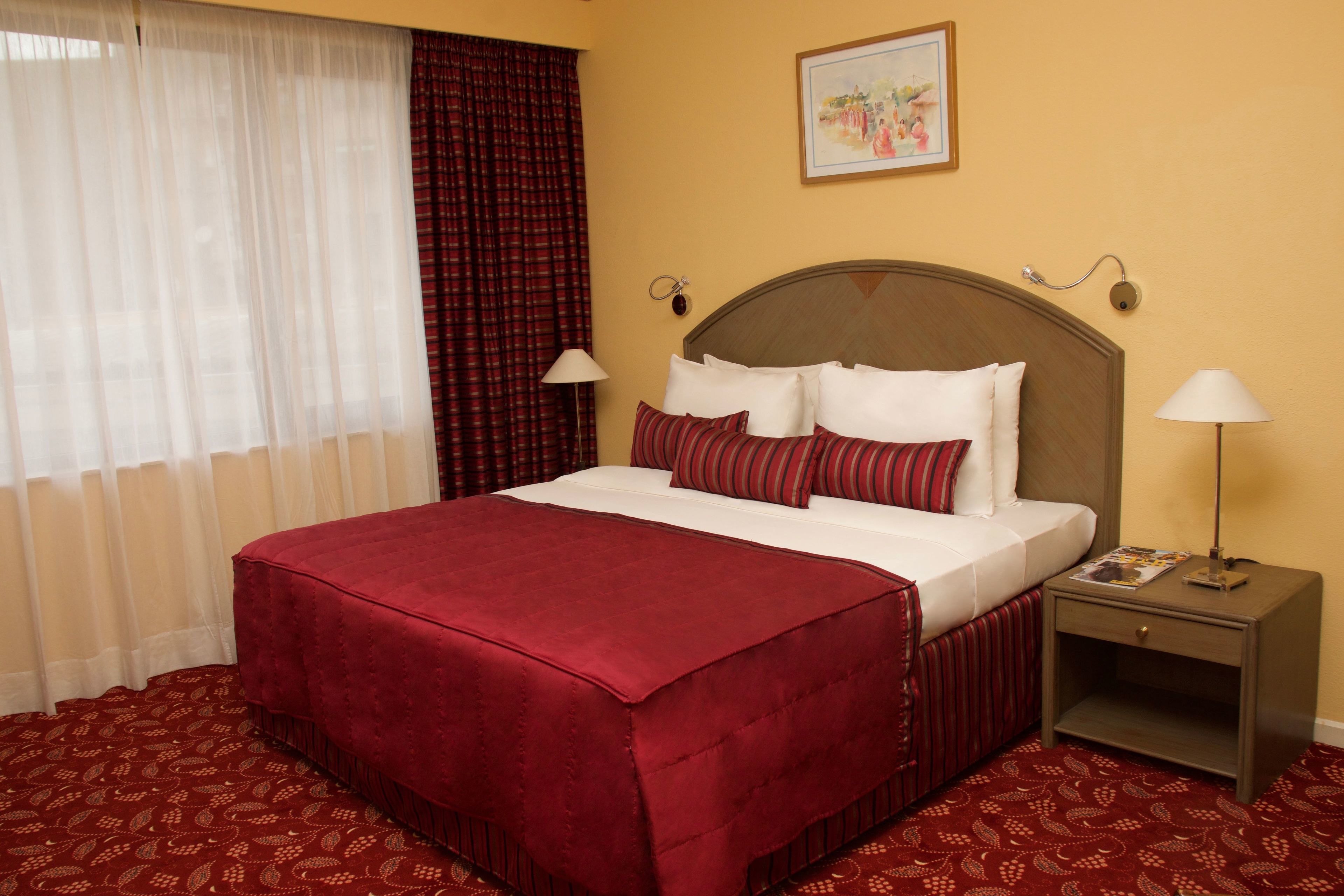 A hotel room with a large bed, two bedside tables, a window with sheer and heavy curtains, and a painting on the wall.