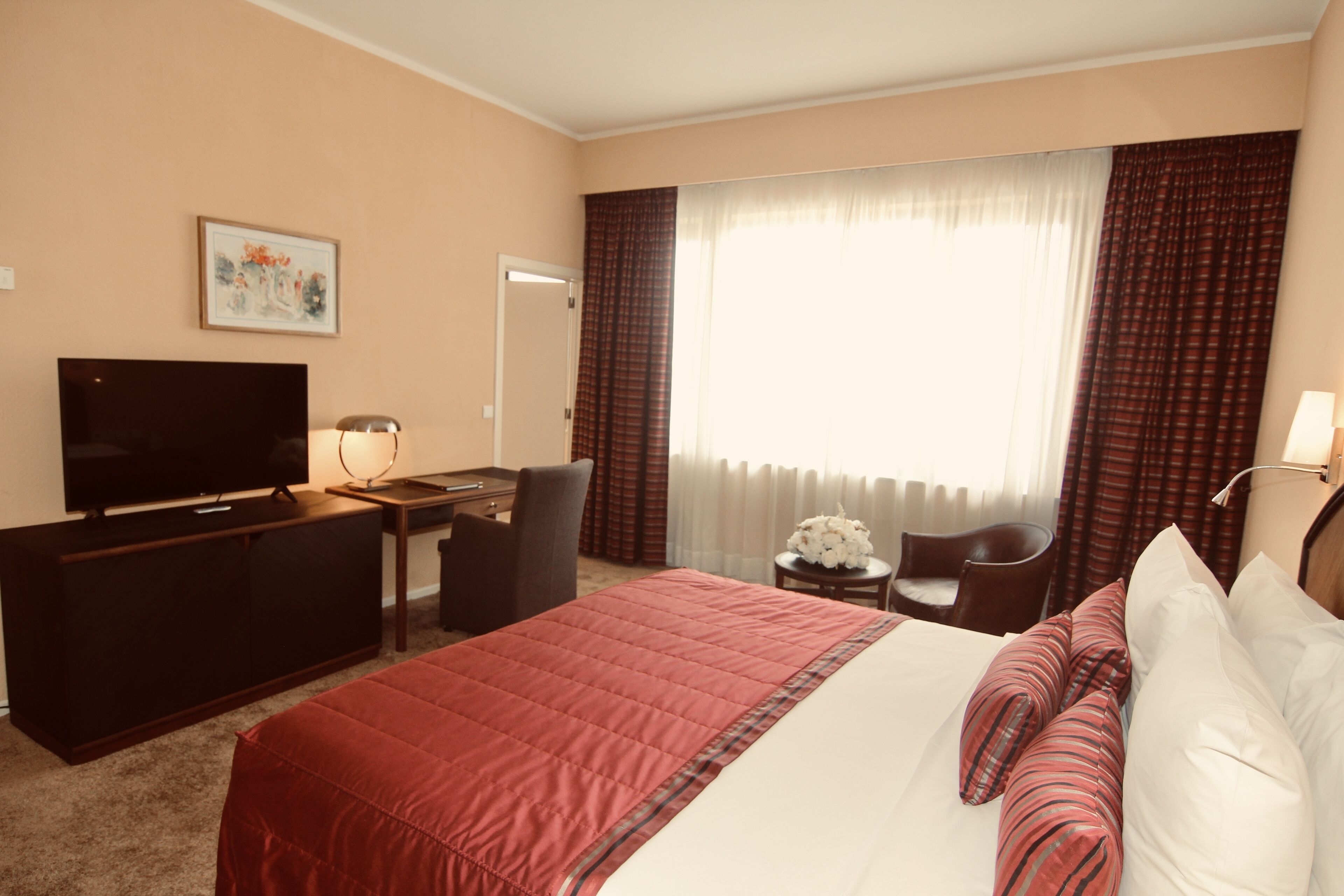 A hotel room with a bed, a desk with a TV, a chair, a small table with a lamp, and a window with curtains.