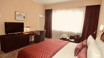 A hotel room with a bed, a desk with a TV, a chair, a small table with a lamp, and a window with curtains.