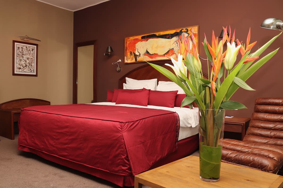 A hotel room with a large bed, a wooden nightstand, a desk, a brown leather chair, and a vase with flowers.