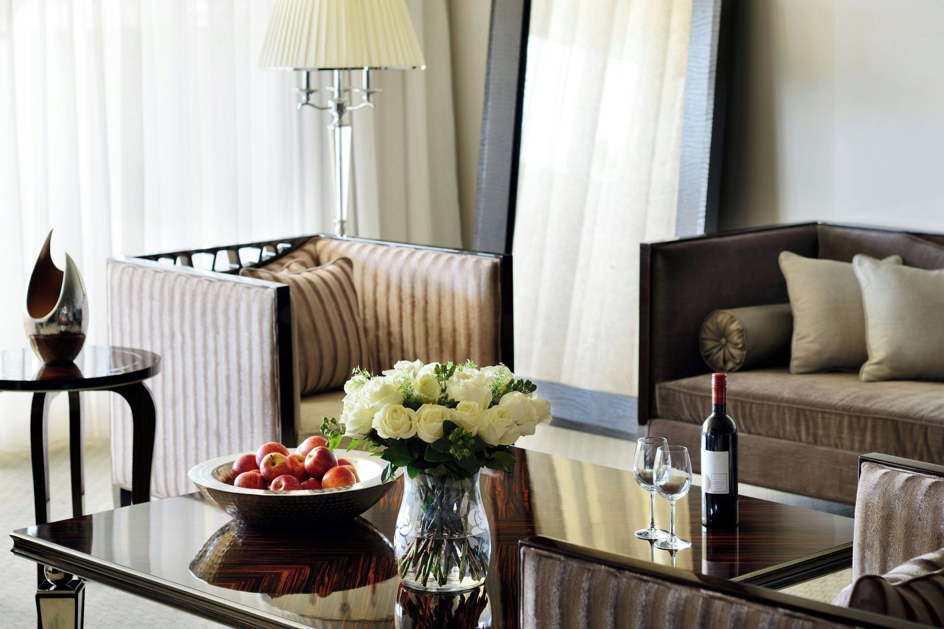 A hotel room with a dining table, a vase with flowers, a bowl of fruit, a bottle of wine, and a sofa.