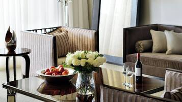Habitación de hotel con una mesa de comedor, un florero con flores, un frutero con fruta, una botella de vino y un sofá.
