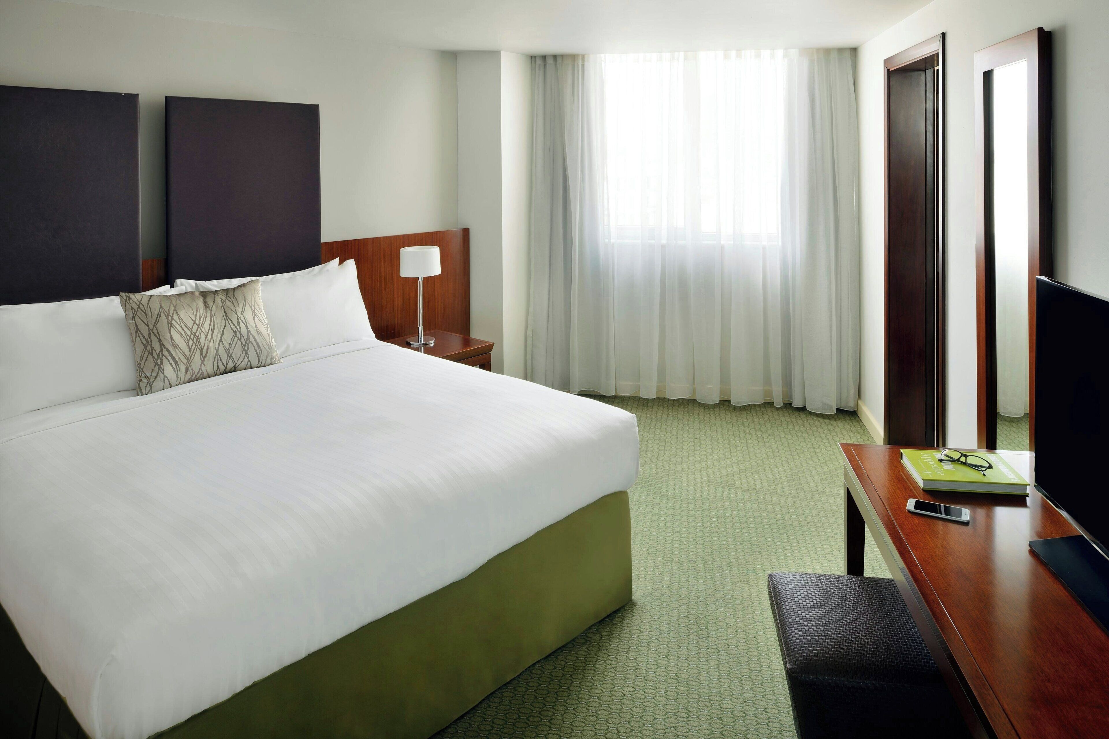 A hotel room with a large bed, a bedside table with a lamp, a desk with a chair, and a TV.