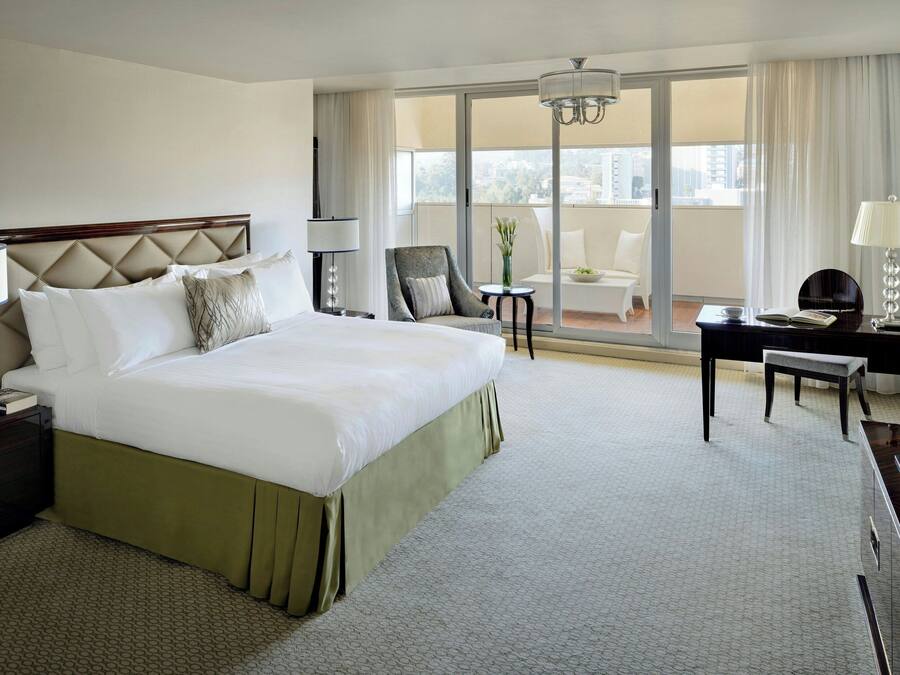 A hotel room with a large bed, a desk, a chair, a lamp, and a view of the city through the window.