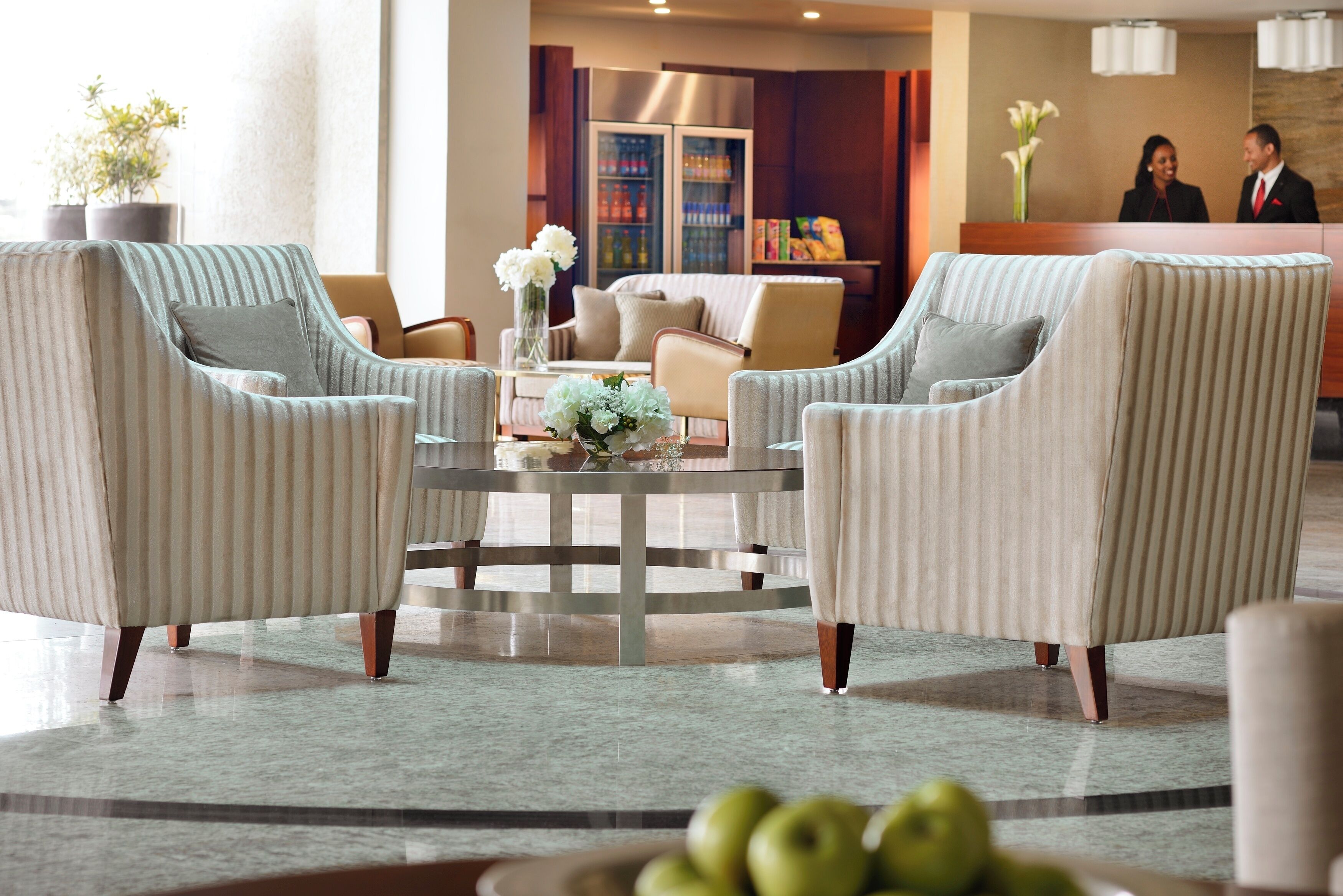 Eine Lobby mit gestreiften Sesseln, einem runden Tisch mit Blumenvase und einem Empfangsbereich mit Kühlschrank und Bücherregalen im Hintergrund.