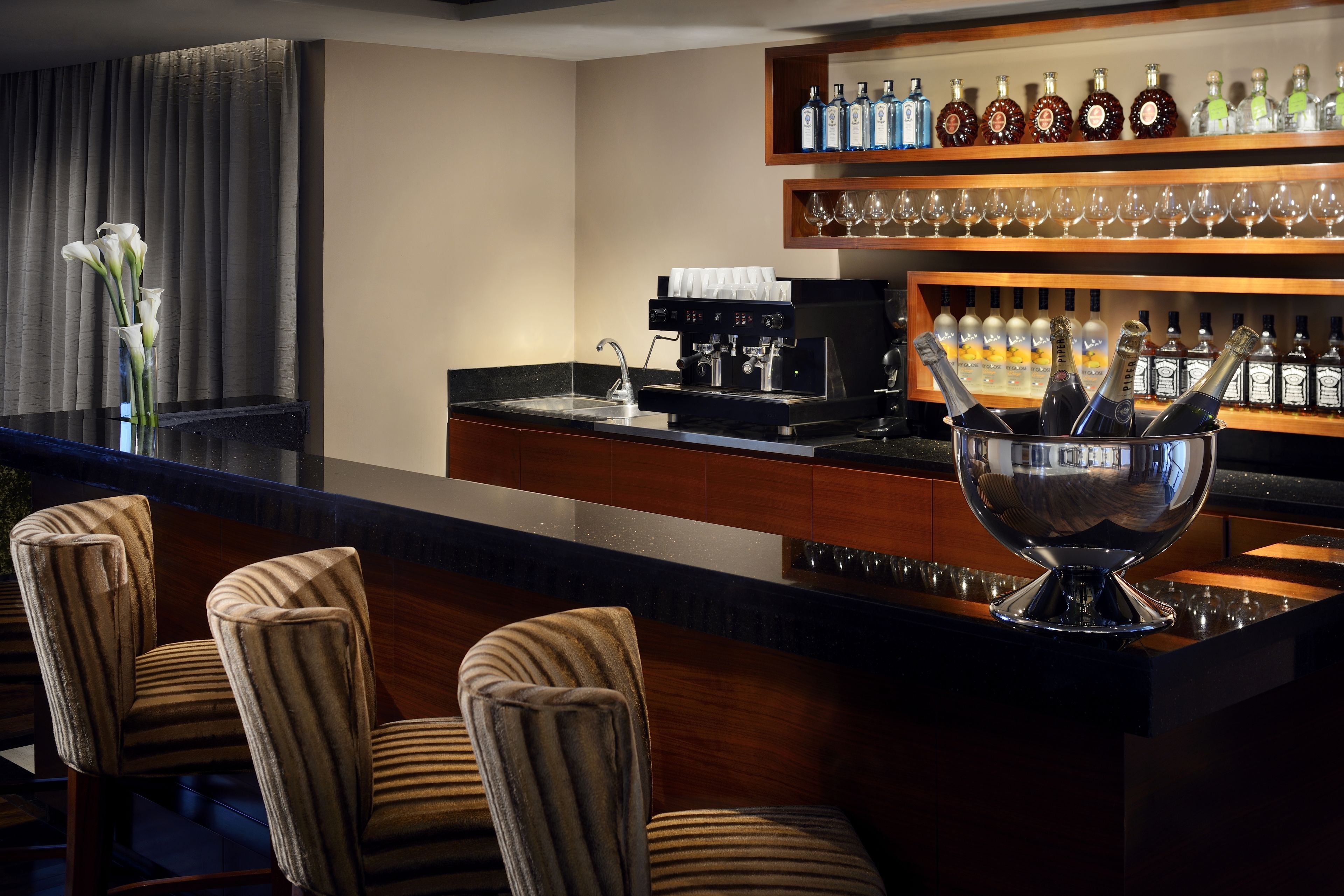 A bar area with a counter, bar stools, a coffee machine, and shelves with bottles and glasses.